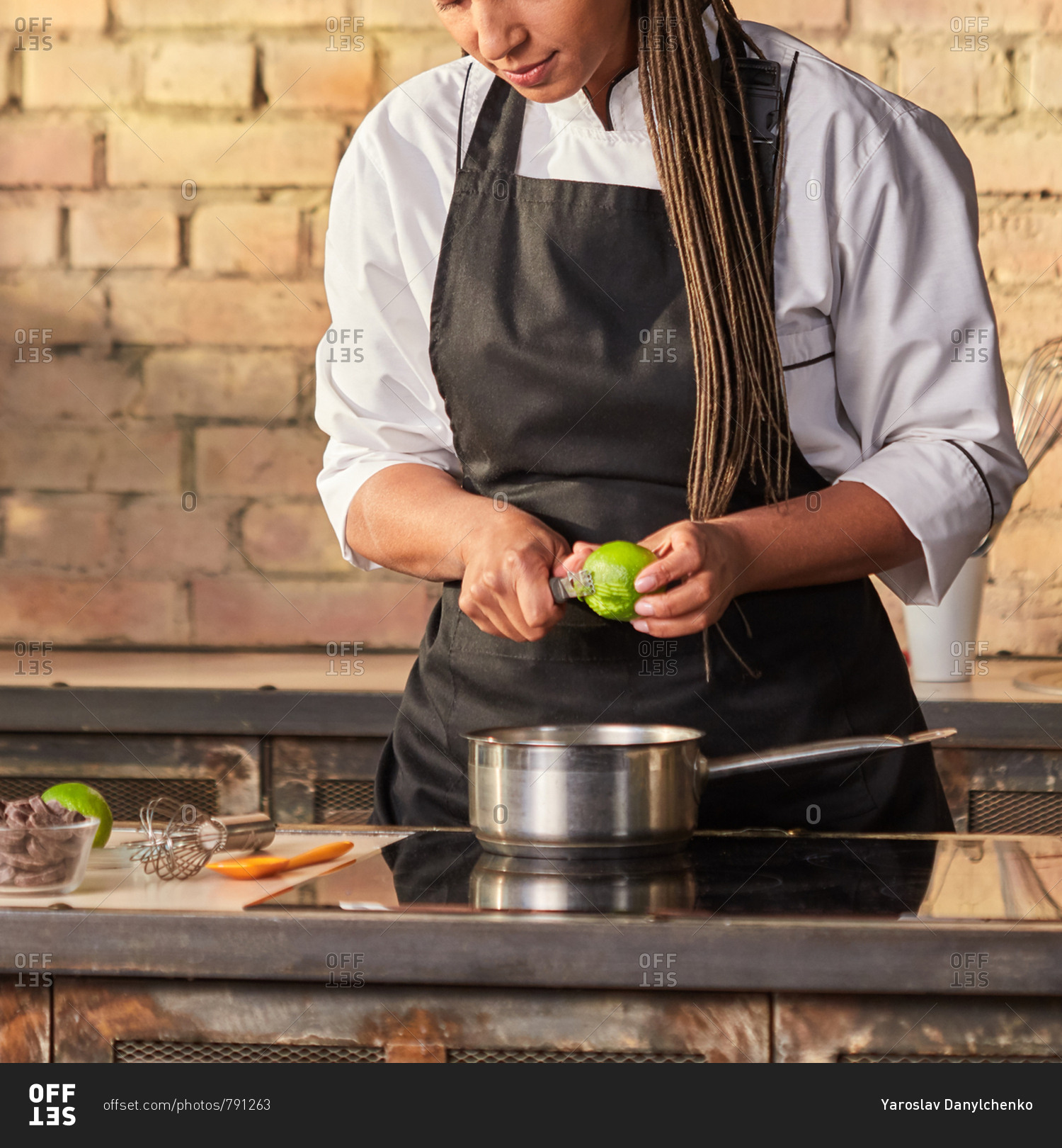 Woman baker cuts lime peel for cooking homemade desserts, green mint on the kitchen table on a brick wall background. Fresh natural ingredients for desserts. Step by step dessert making process.