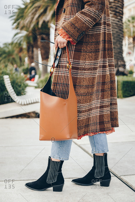Fashion blogger outfit details. Fashionable woman wearing checked oversized coat, red dot dress, regular blue jeans, hobo bag, sunglasses and black suede ankle boots. Mediterranean city surrounding.