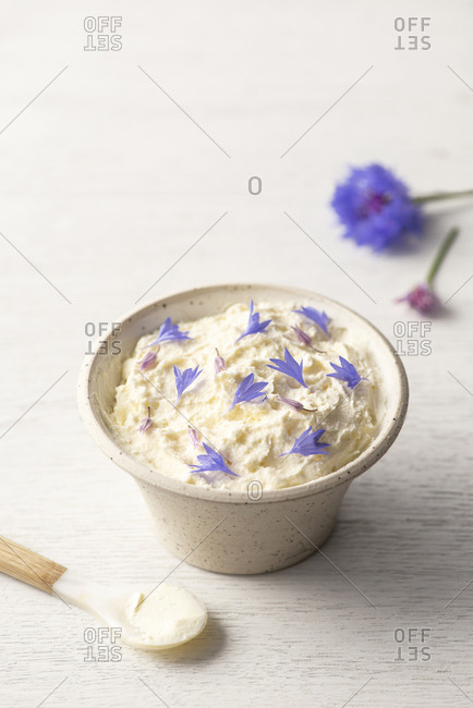 Homemade ricotta with edible flowers in a bowl on a white wooden surface with a spoon.