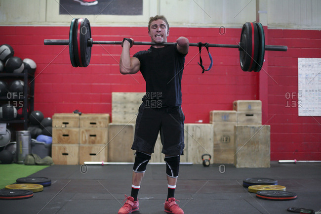 Confident male adaptive athlete lifting barbell while standing against wall in gym