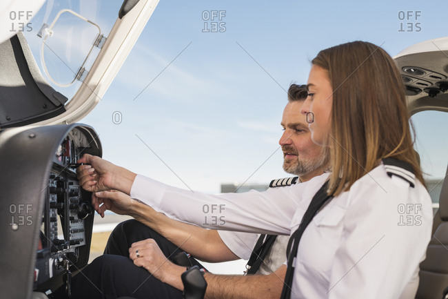 Side view of male pilot teaching female trainee to operate control panel in airplane against blue sky at airport