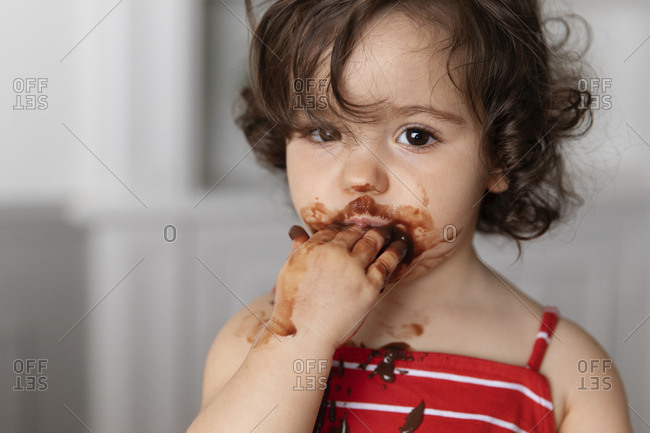 Close-up portrait of cute baby girl with messy face eating chocolate while sitting against wall at home