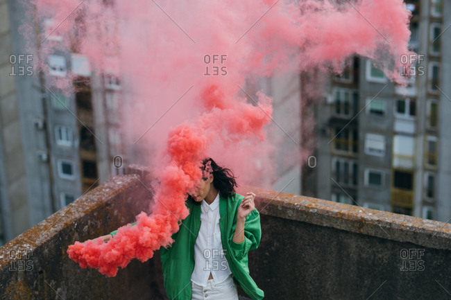 Young woman wearing green jacket holding pink smoke boom on rooftop.