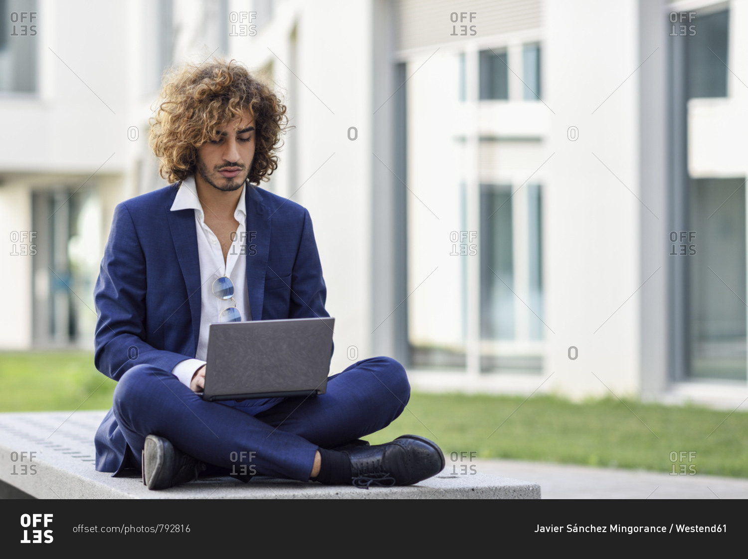 Portrait of young businessman with curly hair wearing blue suit sitting on bench outdoors using laptop