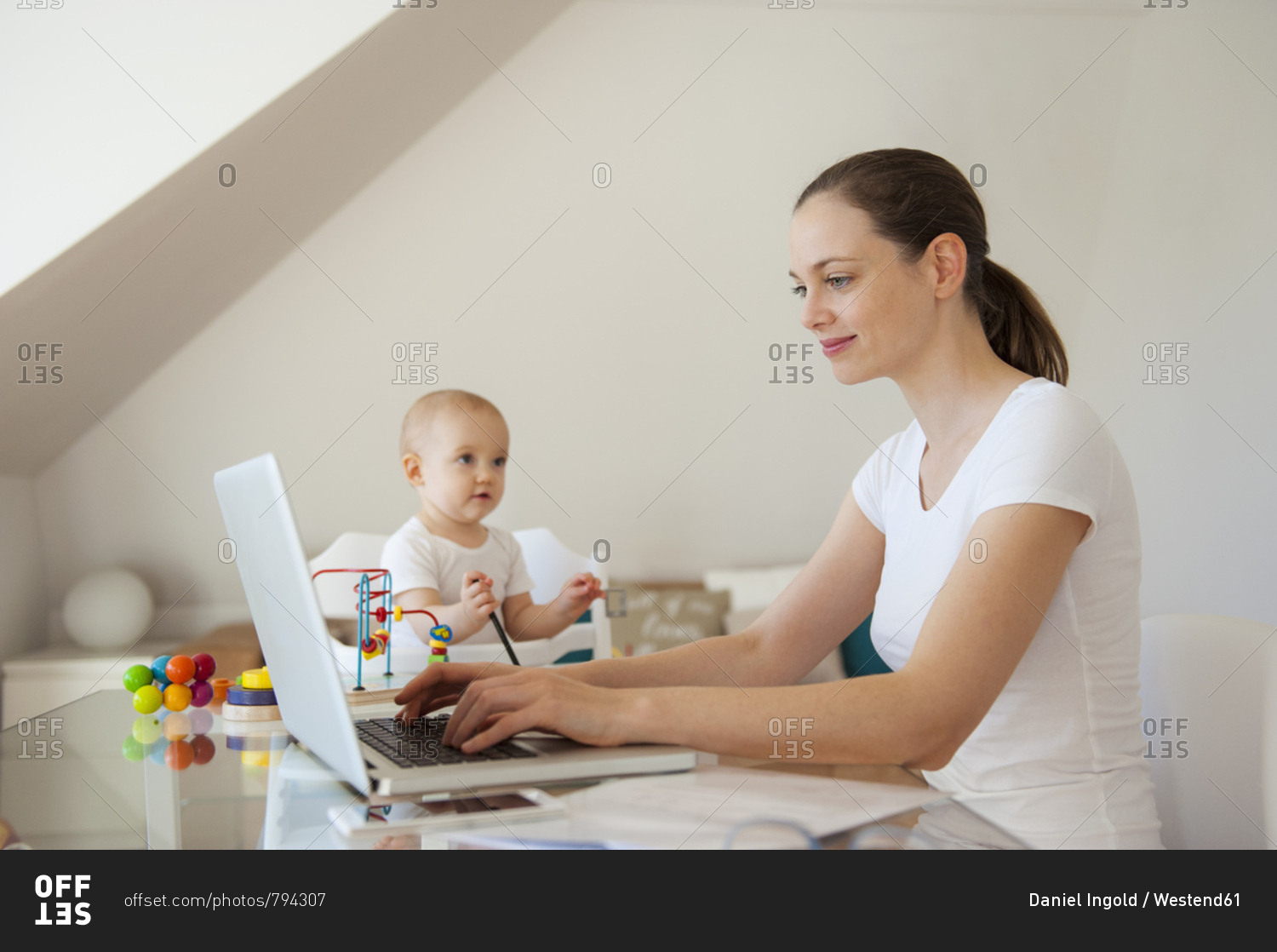Smiling mother using laptop and little daughter playing at table at home