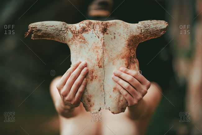 Naked woman holding animal skull covering face while standing in wild nature near rocky cliff