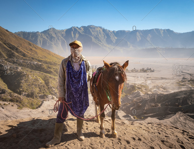 Java Island, Indonesia - August, 14 2015: Ethnic person with scarf on face looking at camera while standing near lovely horse not far from Mount Bromo on sunny day