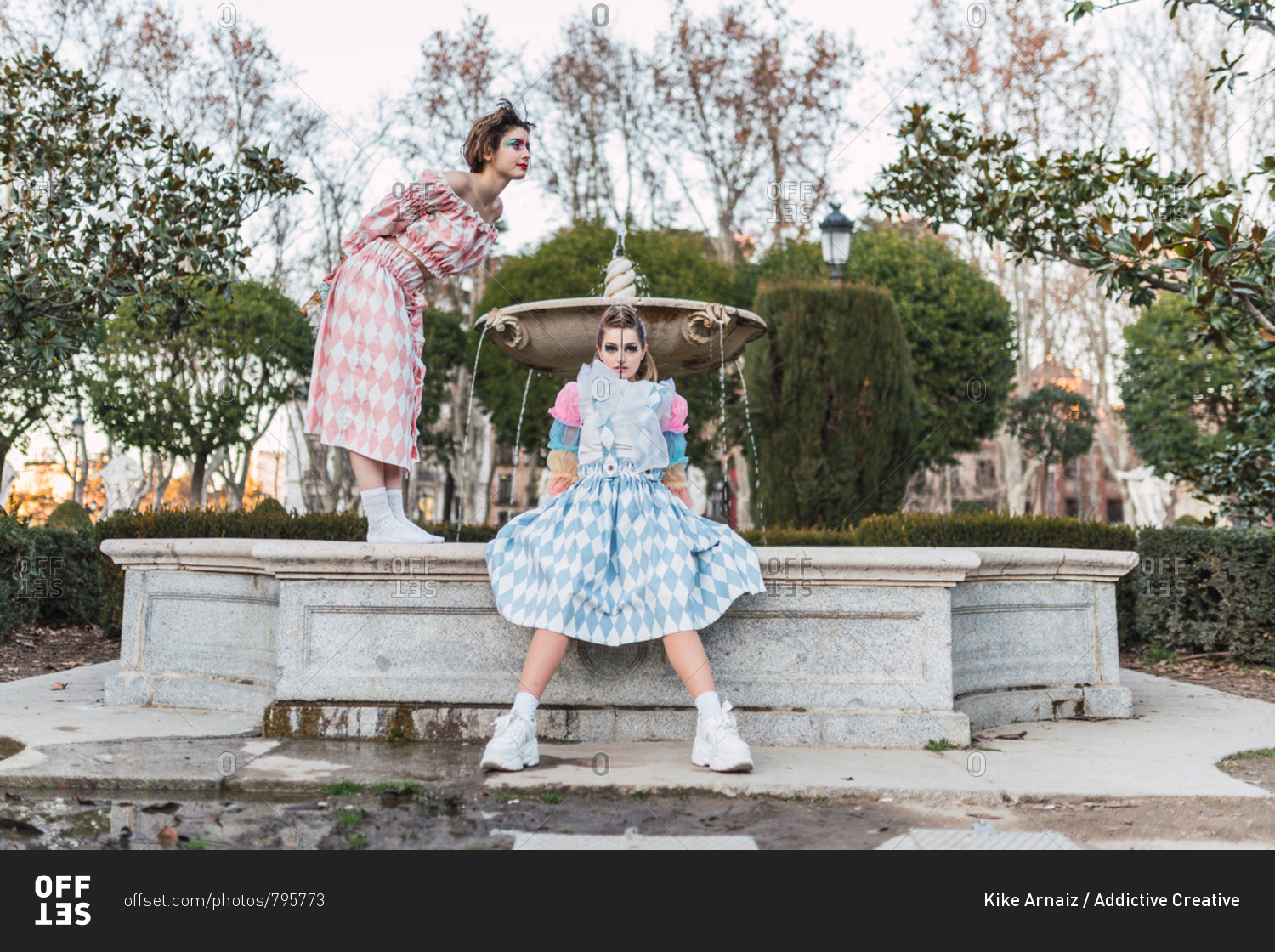 Two young women in theatrical costumes acting near old fountain in beautiful park
