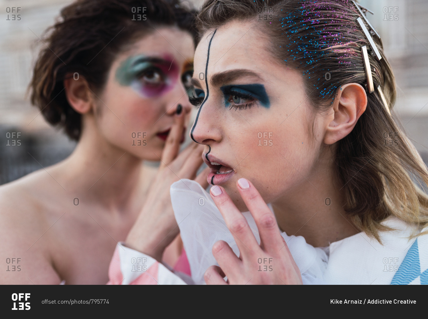 Two young women with theatrical makeup and unusual clothes sharing secret while performing on city street
