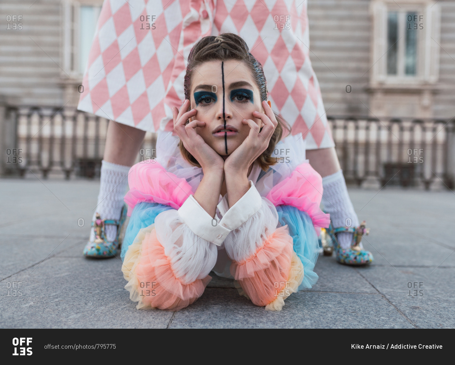 Young woman with theatrical makeup and fancy clothes looking at camera while lying on ground between legs of female street performer