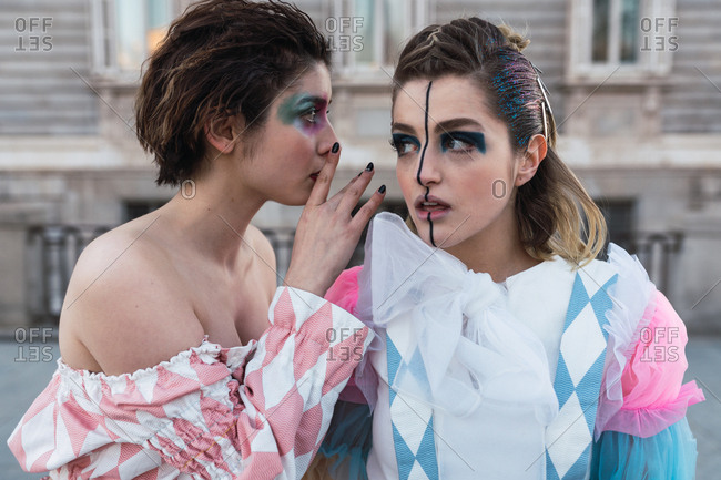 Two young women with theatrical makeup and unusual clothes sharing secret while performing on city street