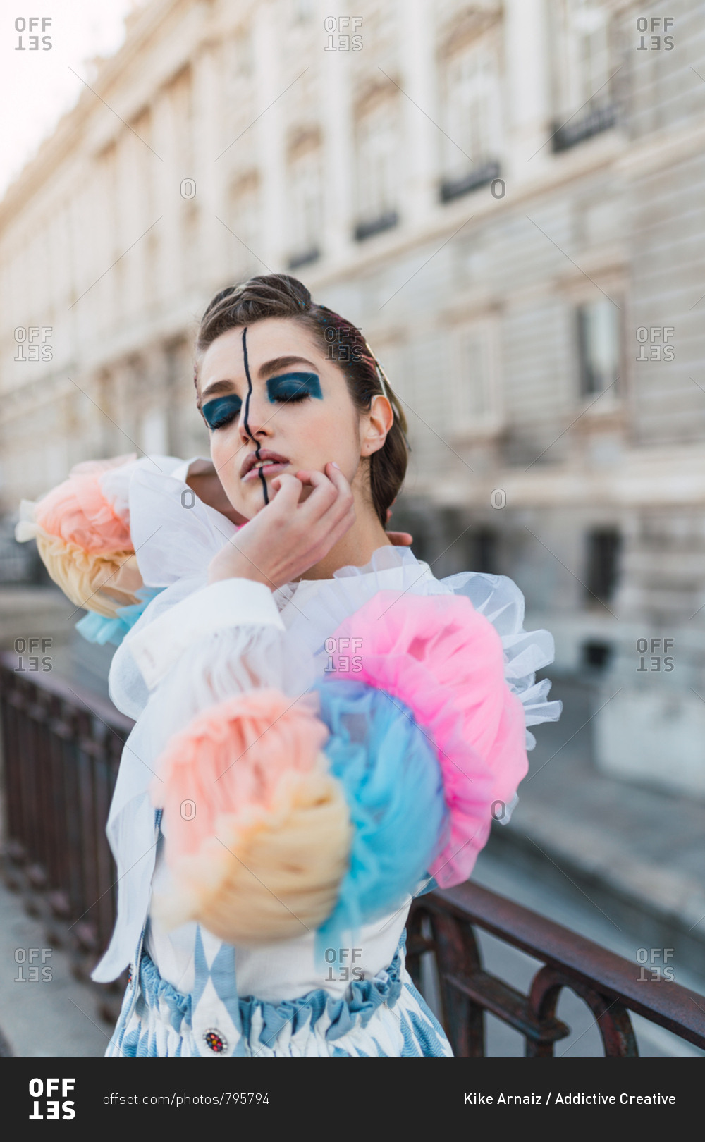 Beautiful young lady with theatrical makeup keeping eyes closed and touching face while standing near fence on blurred background of city street