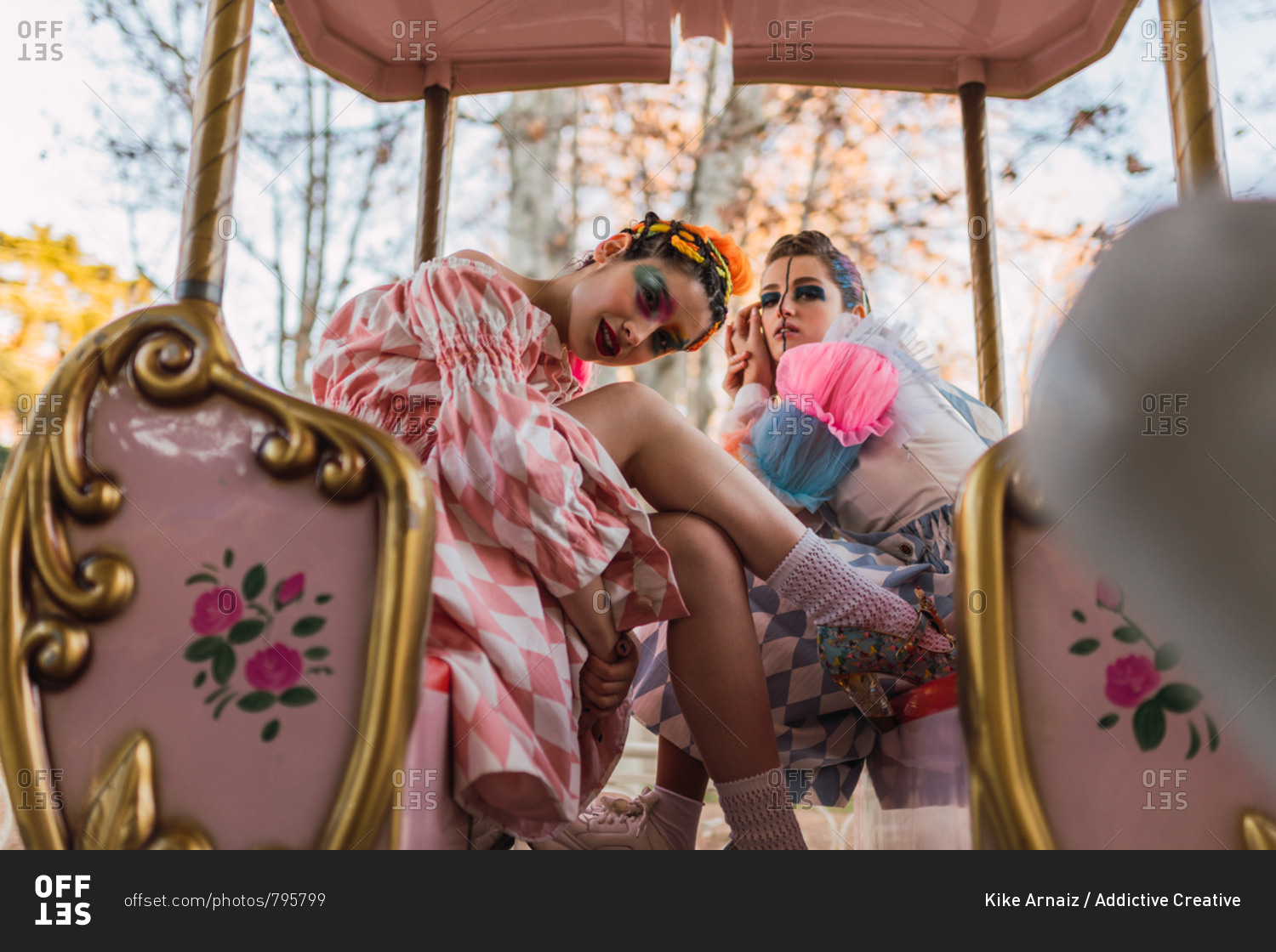 Two pretty young women with theatrical makeup and costumes looking at camera while sitting on amazing roundabout in amusement park