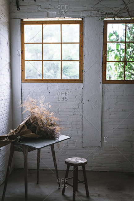 Concept of bouquet of dry coniferous twigs in craft paper on table near stool in grey murk room with brick walls