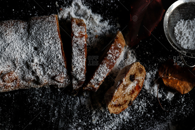 Slices of yummy fresh Stollen lying on dark tabletop near small sieve with powdered sugar and dry autumn leaves