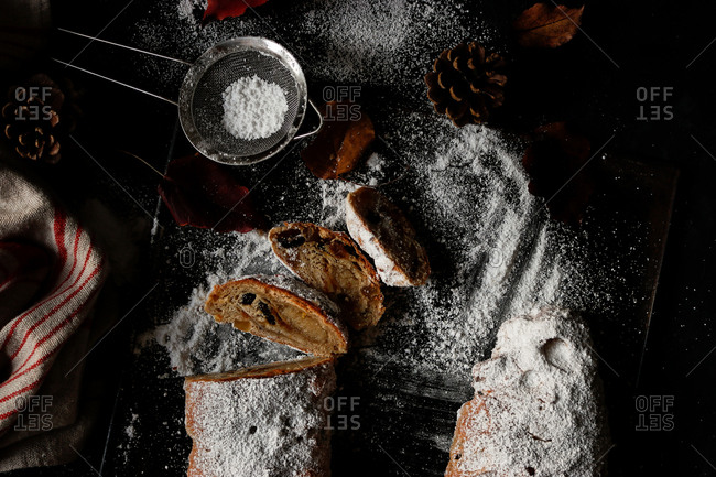 Slices of yummy fresh Stollen lying on dark tabletop near small sieve with powdered sugar and dry autumn leaves