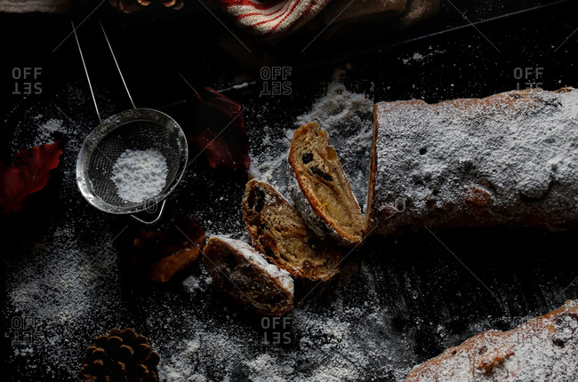 Slices of yummy fresh Stollen lying on dark tabletop near small sieve with powdered sugar and dry autumn leaves