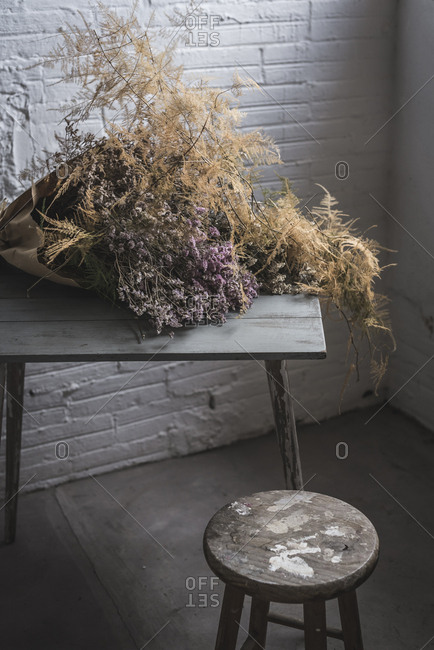 Concept of bouquet of dry coniferous twigs in craft paper on table near stool in grey murk room with brick walls