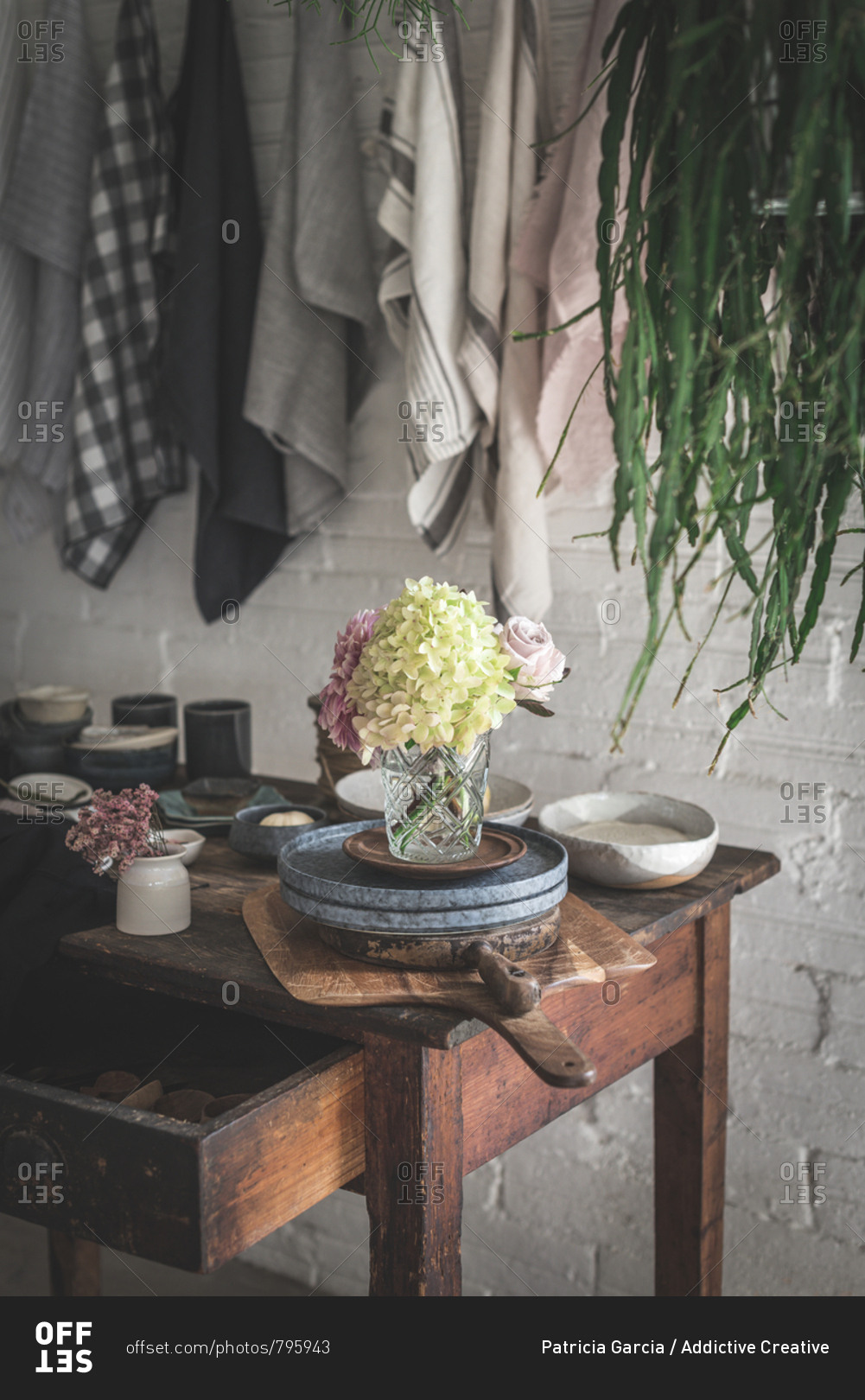 Wooden table with bunch of fresh pink chrysanthemums and white hydrangea in vase between frying pan and kitchenware near dish cloths hanging on twist with pins