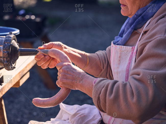 Unrecognizable elderly woman using metal machine to fill animal bowel with minced meat
