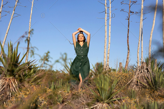 Pretty young model in green dress holding hands over head while standing near thin exotic trees and bushes on sunny day in nature