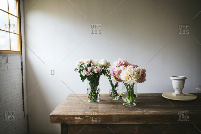 Wooden table with kitchenware and bouquets of fresh blooms in vases with water near white wall