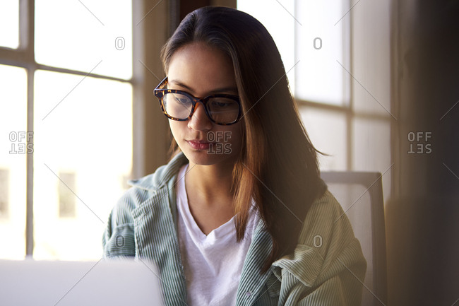 Young female creative using a laptop computer by a window in an office, close up, selective focus