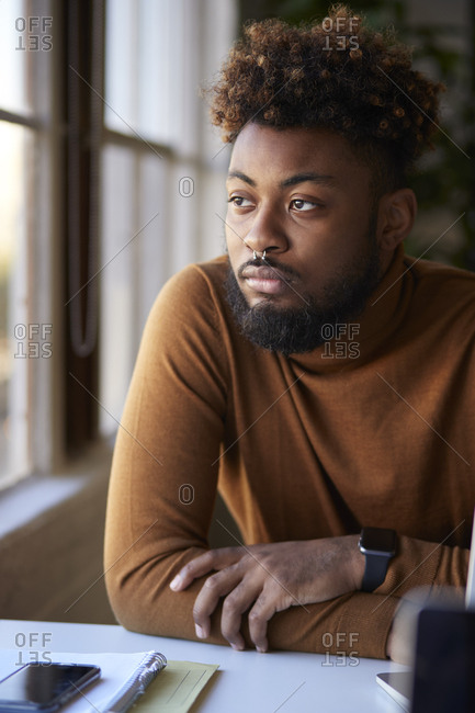 Millennial male creative sitting at a desk in an office looking out of the window, vertical close up