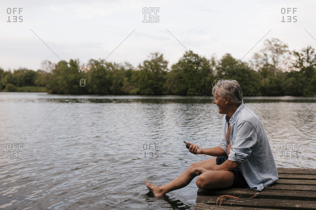 Happy senior man sitting on jetty at a lake with cell phone