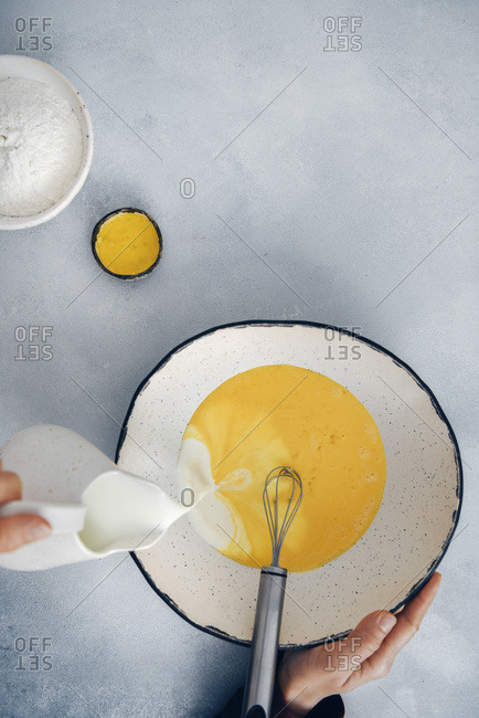 Woman pouring milk into beaten eggs in a large ceramic mixing bowl.