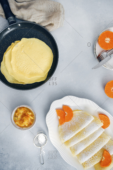 Gluten free crepes in a non stick pan and in a white oval plate garnished with orange slices and powdered sugar, accompanied by orange jam.