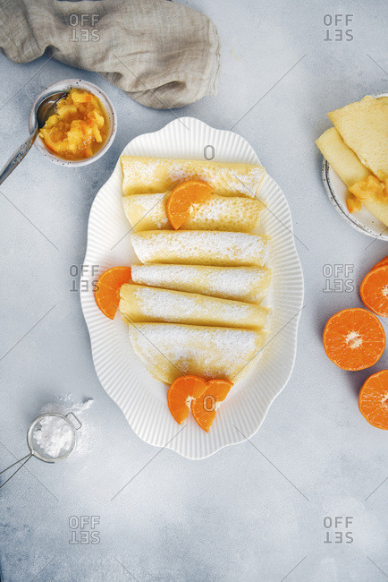 Gluten free crepes served in a white oval plate, garnished with orange slices and powdered sugar, accompanied by orange jam, mini strainer and halved oranges.