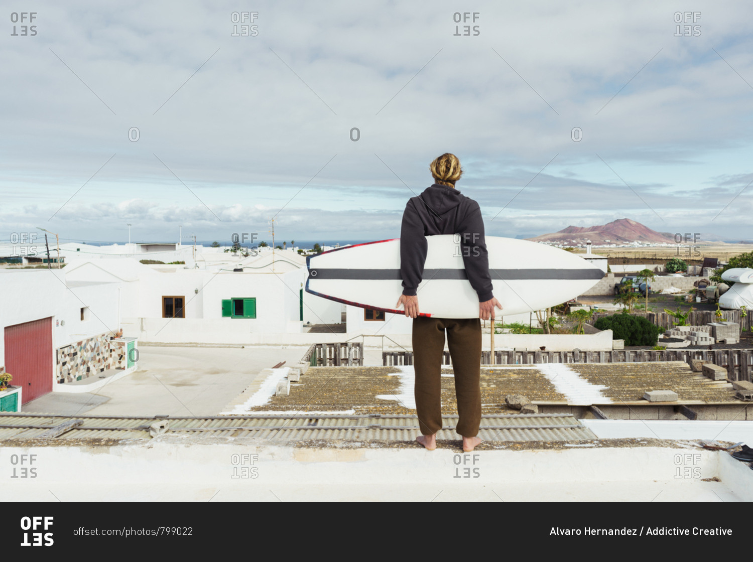 Back view of young guy in sweater standing with craft surf board on roof and looking at houses and stone hill
