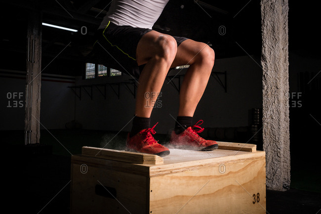 Crop athletic young guy in sportswear jumping on wooden box between chalk dust in gym