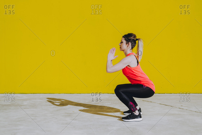 Young lady in sportswear doing squad exercises near yellow wall