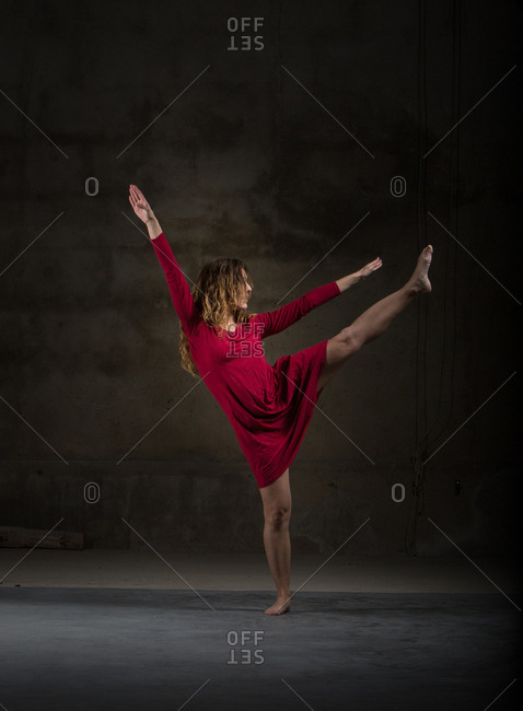 Young slim ballerina in red dress upping leg and hands in dark room