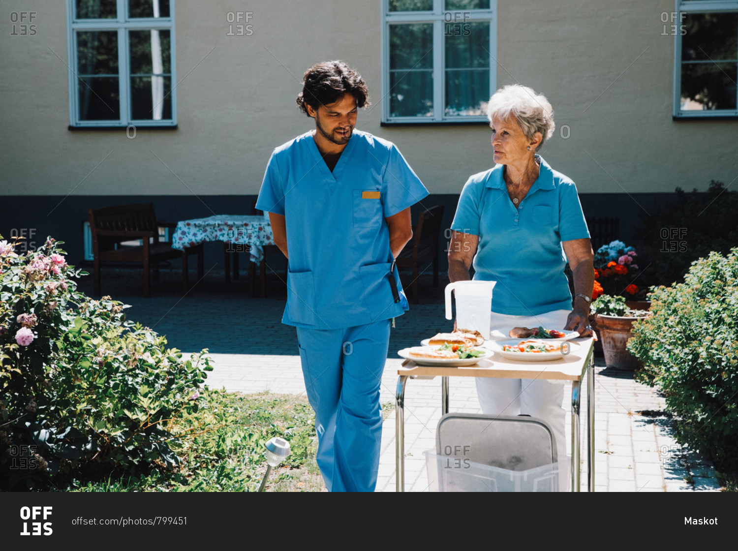 Male nurse walking with senior woman pushing food cart against nursing home
