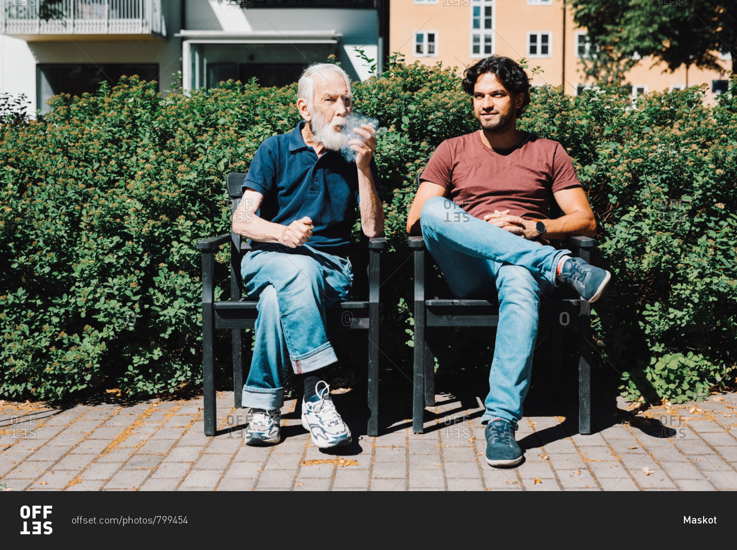 Full length of retired senior man smoking pipe while sitting with male caretaker against nursing home