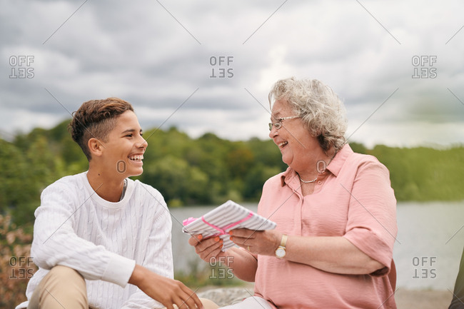 Cheerful grandmother giving gift to grandson in park on lakeshore during picnic
