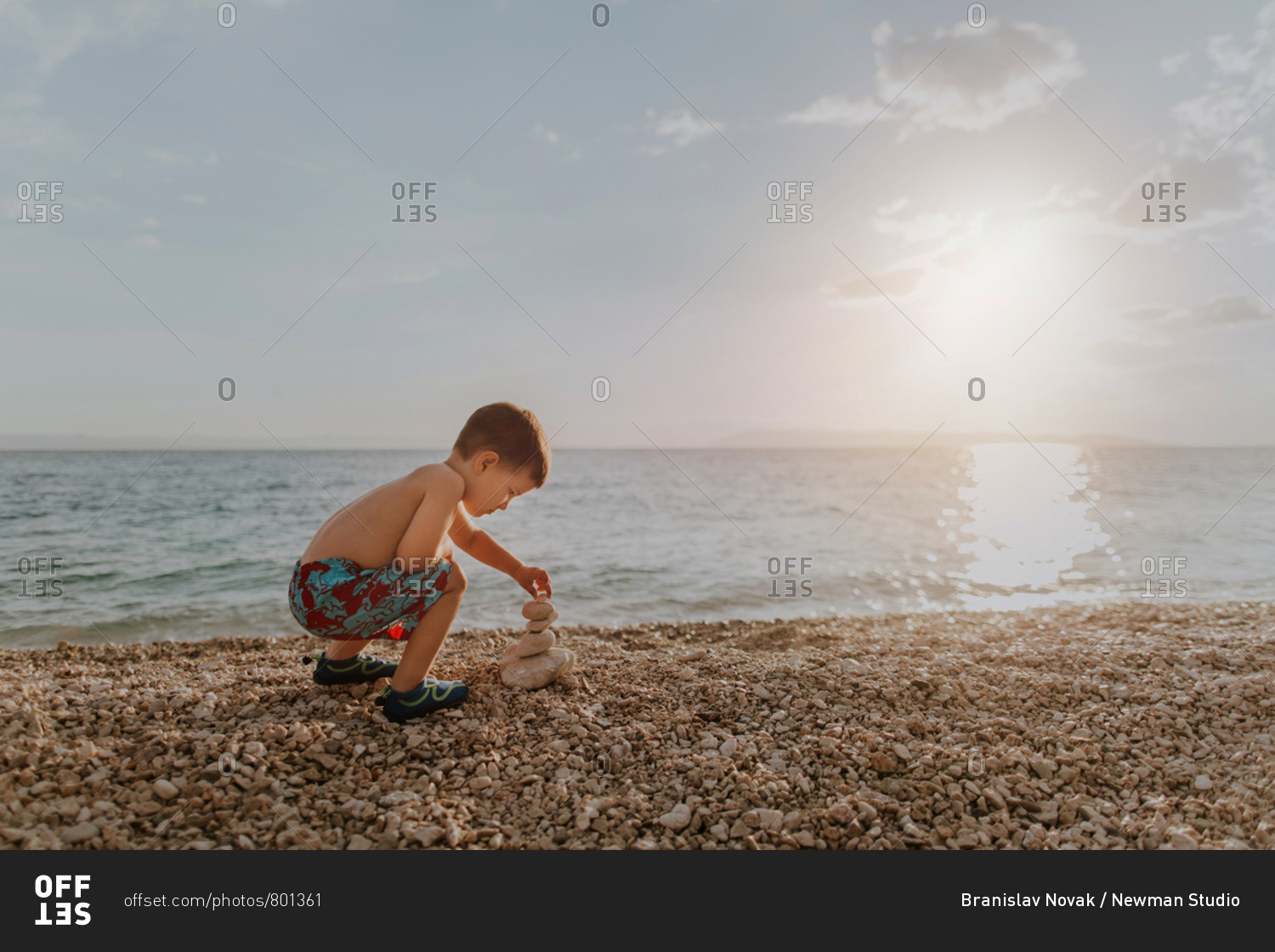 Child playing with stones on beach on his own at sunset. Young boy wearing summer shorts stacking pebbles on beach in the evening.