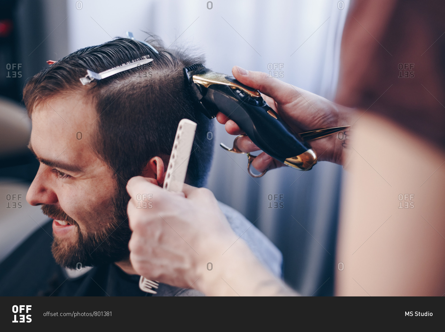 Young handsome man getting trendy haircut at barber shop. Male hairstylist shaving and trimming clients hair using an electric trimmer and comb.
