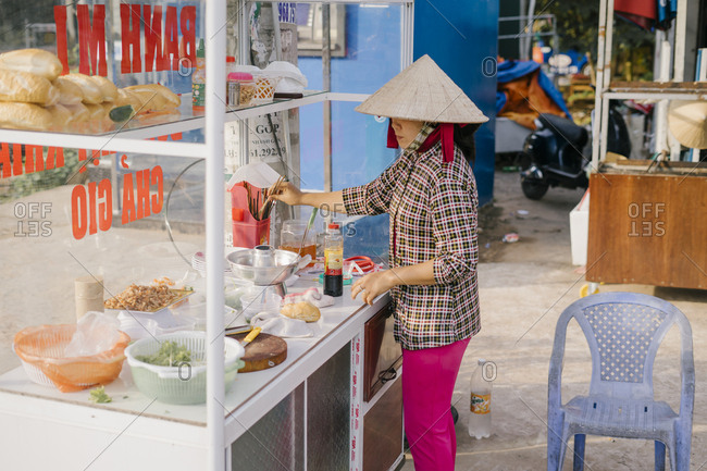 Phu Quoc, Vietnam - January 19, 2019: Vietnamese woman in hat prepares street food sandwich bahn me with ham, pate and vegetables.