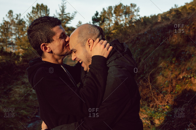 Side view of cheerful homosexual couple embracing on route in forest in sunny day