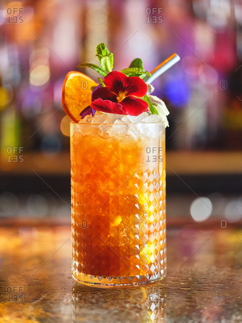 Glass of yummy cocktail decorated with red flower and slices of fresh citrus and placed on counter on blurred background of bar
