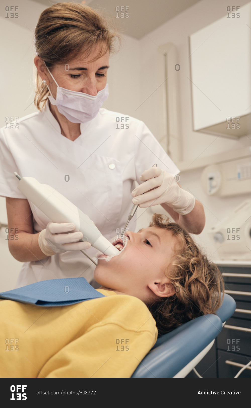 Woman in mask and doctor uniform making scan of teeth of little boy while working in dentist clinic