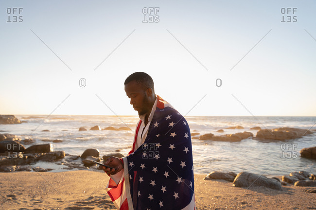 Side view of young African-American man wrapped American flag while using mobile phone on the beach at sunset