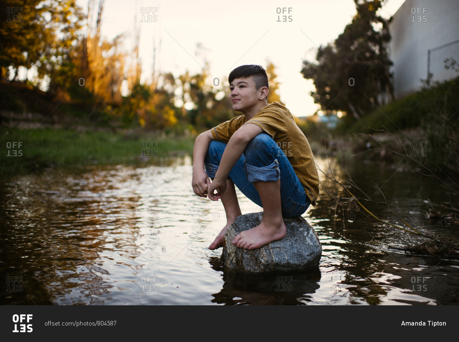 Boy sitting barefoot on rock in a stream stock photo - OFFSET