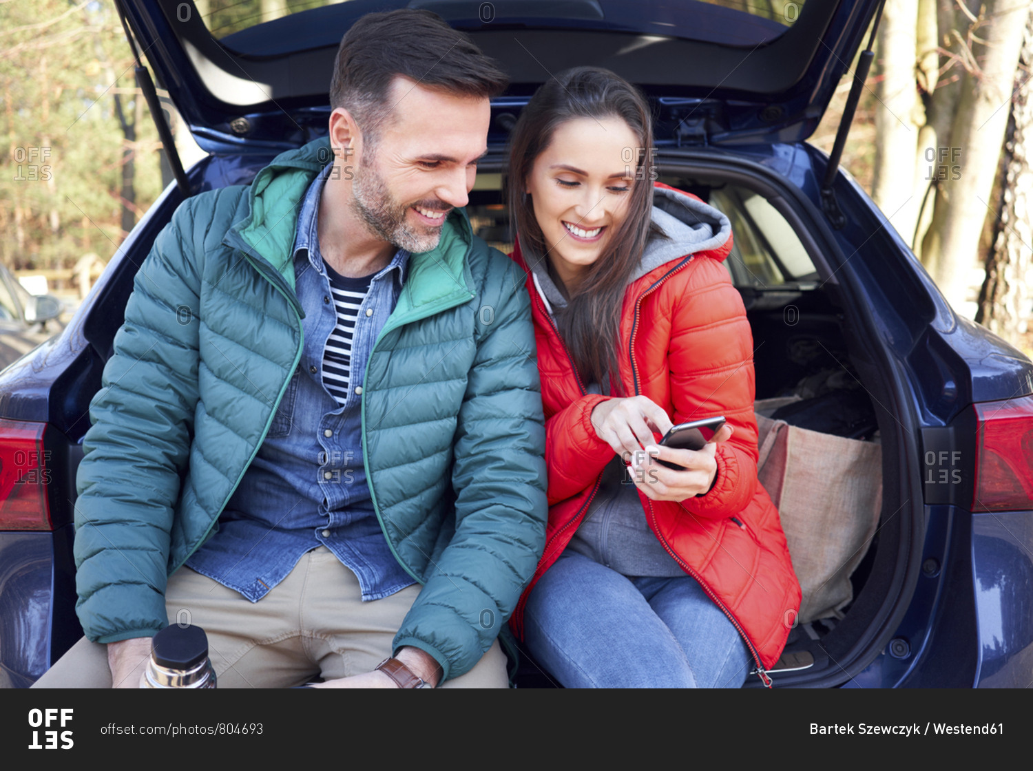 Couple using smartphone while sitting in car trunk during road trip