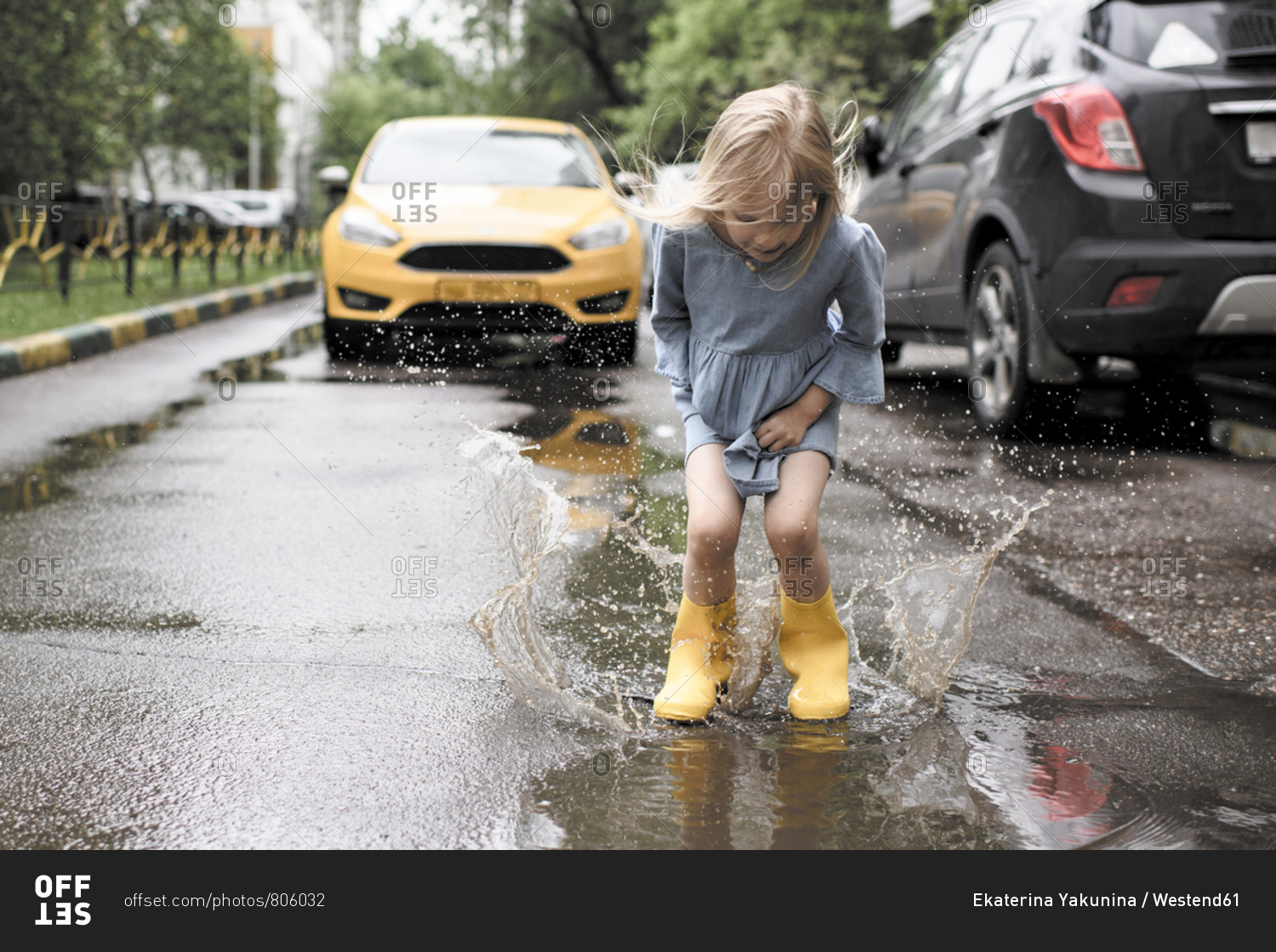 Girl wearing blue dress and rubber boots- jumping in pond on street- yellow car in the background