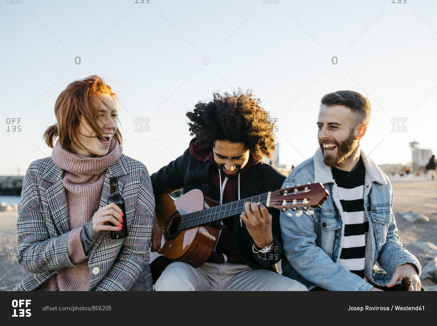 Three happy friends with guitar sitting outdoors at sunset